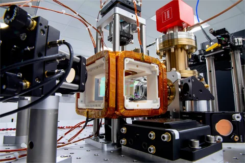Copper coils surround a central glass vacuum cell in QuEra’s Aquila apparatus, with optical mounts, lenses and metal vacuum fittings nearby.