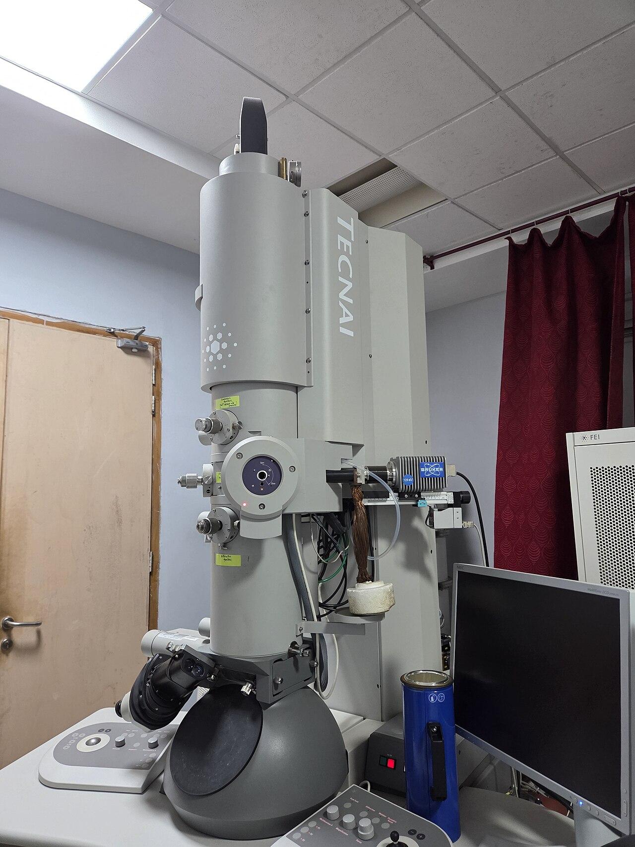 A tall transmission electron microscope with its control equipment in a laboratory.