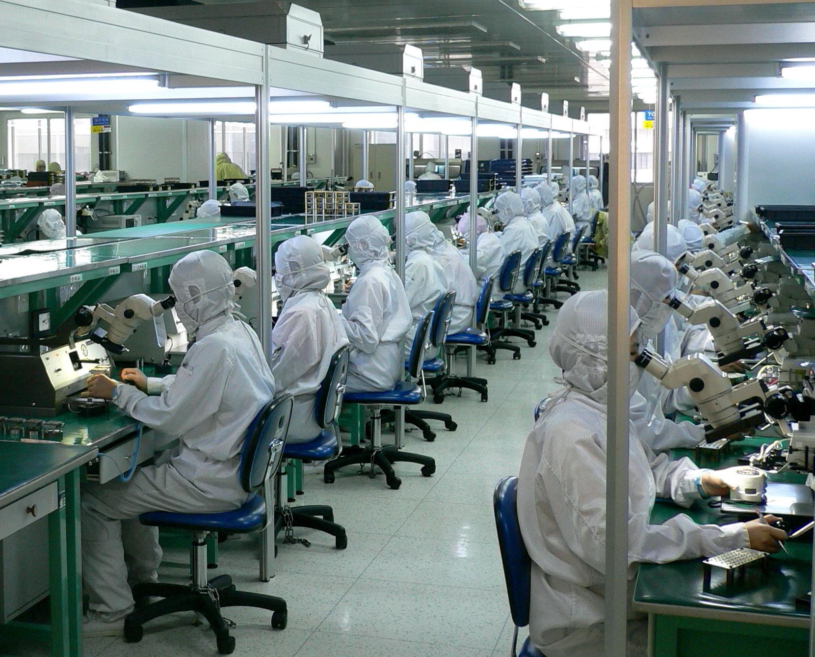 Workers in protective clothing sit along fiber-optic assembly and testing benches in a factory.