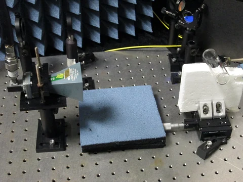 A horn antenna points toward a glass vapor cell among optical components on a laboratory table.
