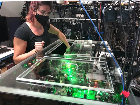 Alejandra Collopy wearing safety glasses and a mask beside an optical bench with green laser light and cables.