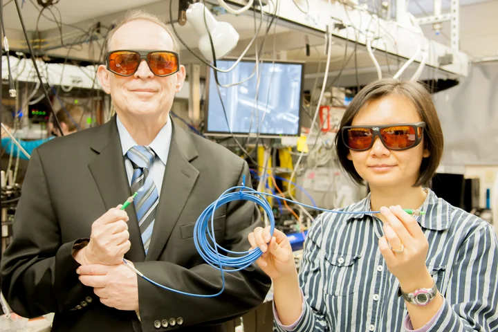 Two researchers wearing laser safety glasses hold opposite ends of a coiled blue fiber cable in a laboratory.