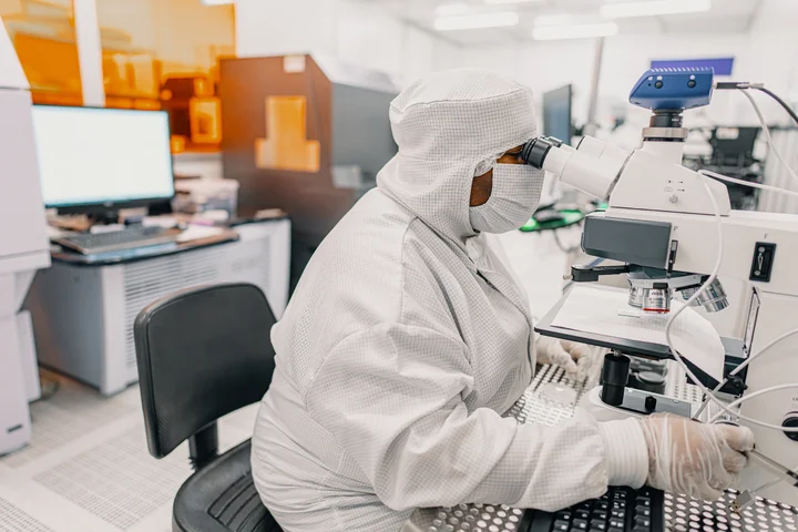 A worker in a white cleanroom suit looks through a microscope at an IQM workbench.