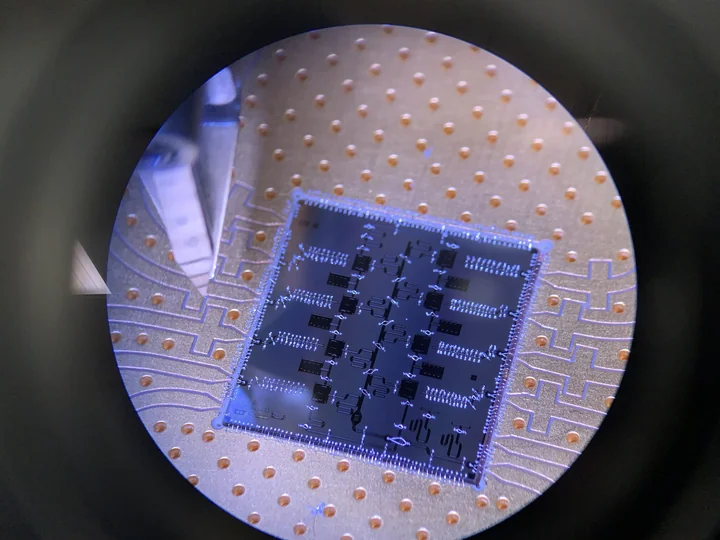 A square blue-purple quantum processor chip with fine patterned structures on a circular gold-colored mount.