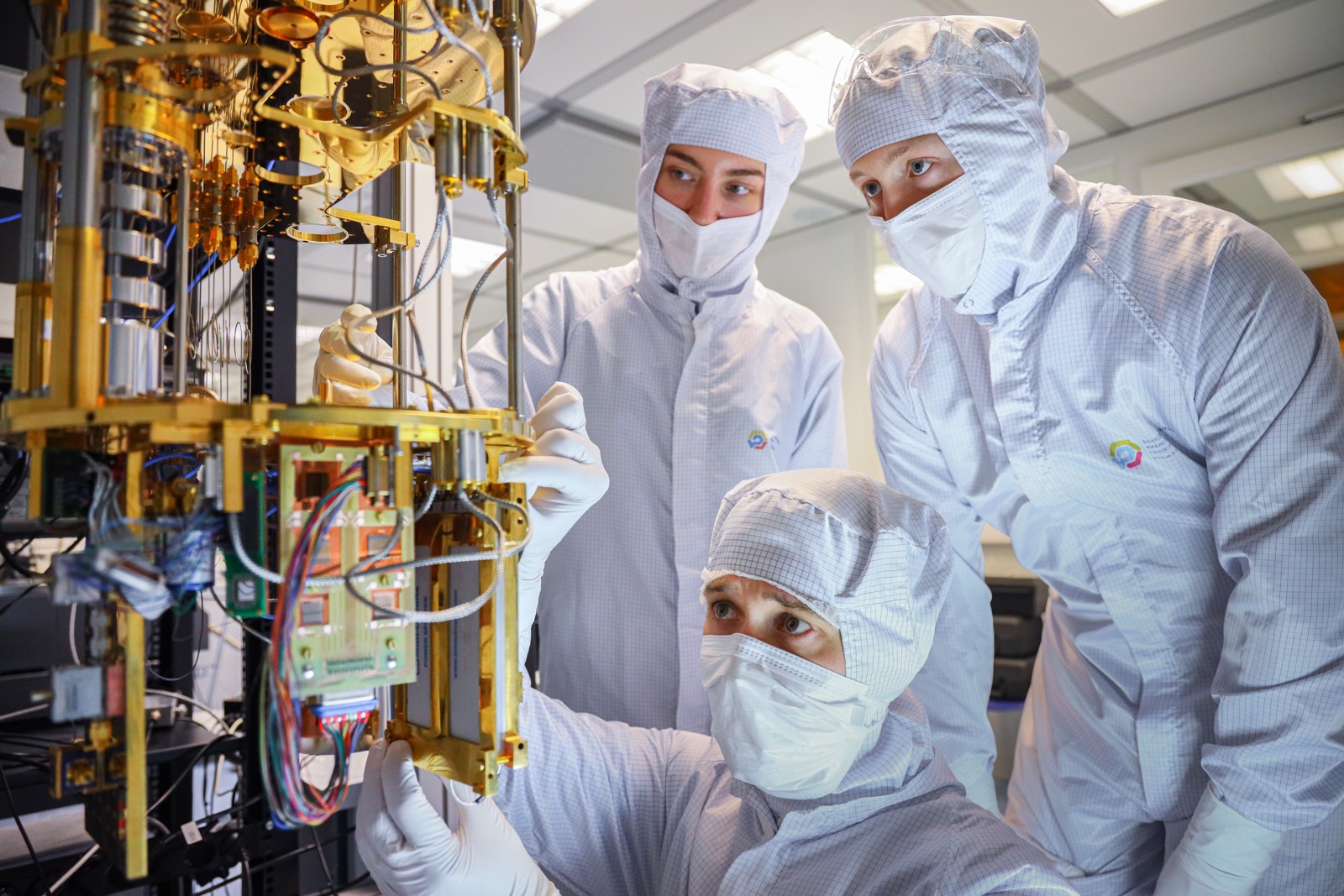 Three FMN Laboratory engineers in cleanroom clothing inspect gold-colored cryogenic stages, microwave components and wiring while assembling superconducting quantum hardware