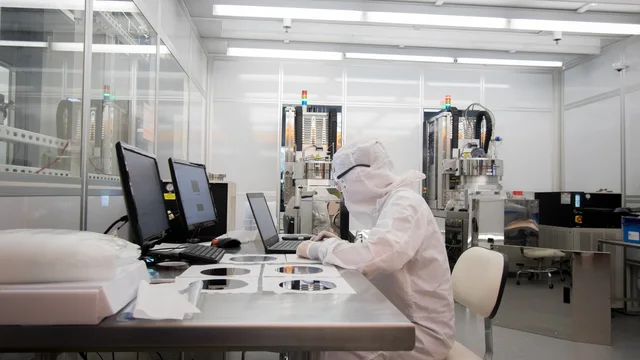 A technician in a white cleanroom suit works beside silicon wafers and fabrication instruments.