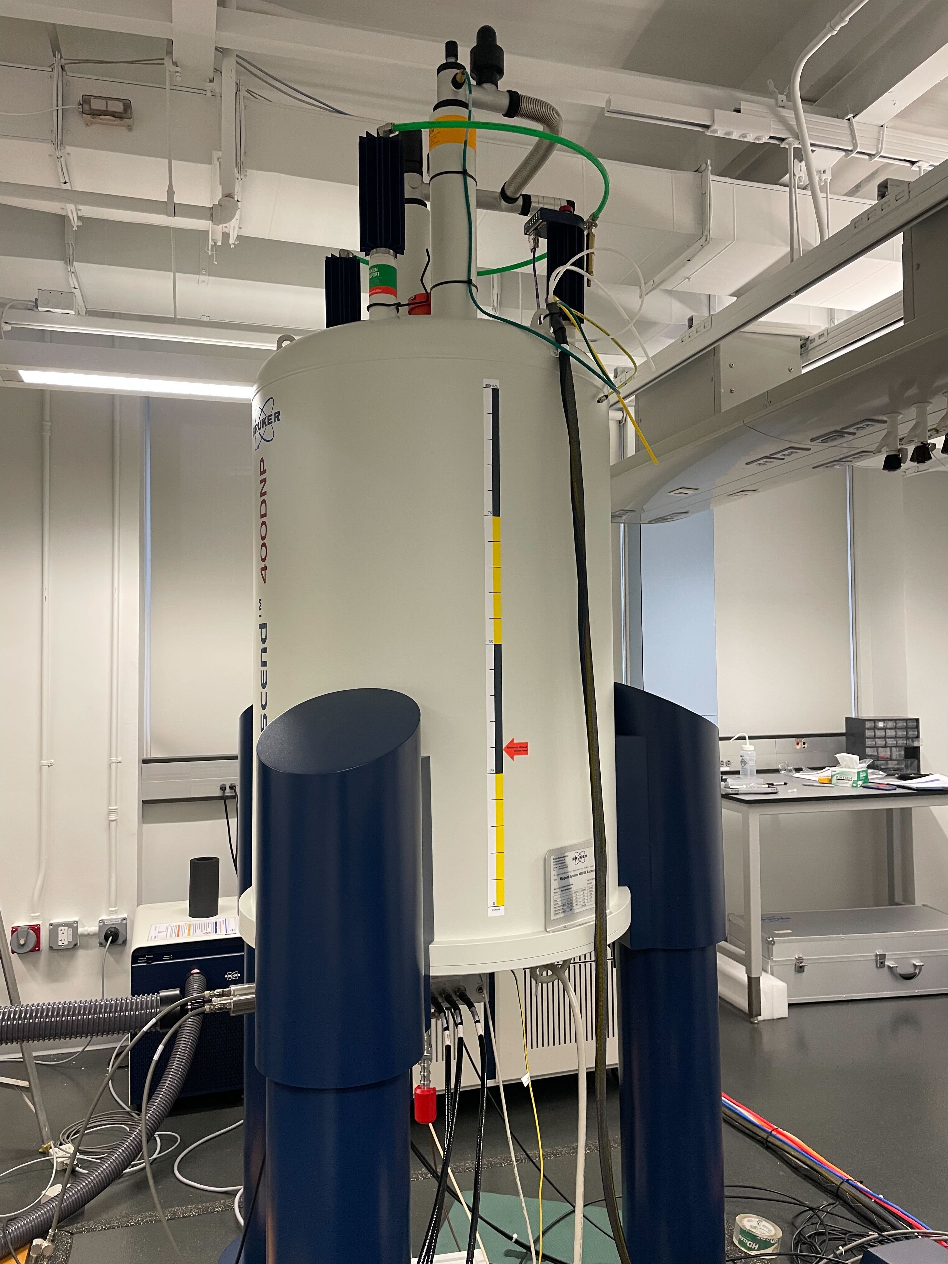 A white cylindrical nuclear magnetic resonance spectrometer with dark supports and connected tubing in a laboratory.