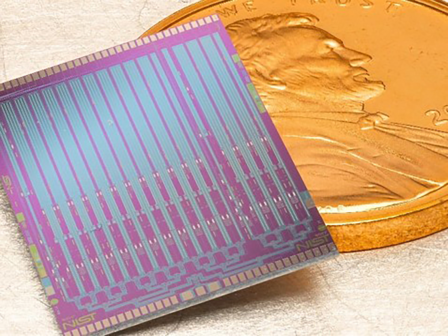A small iridescent NIST Josephson voltage-standard chip beside a U.S. penny for scale.