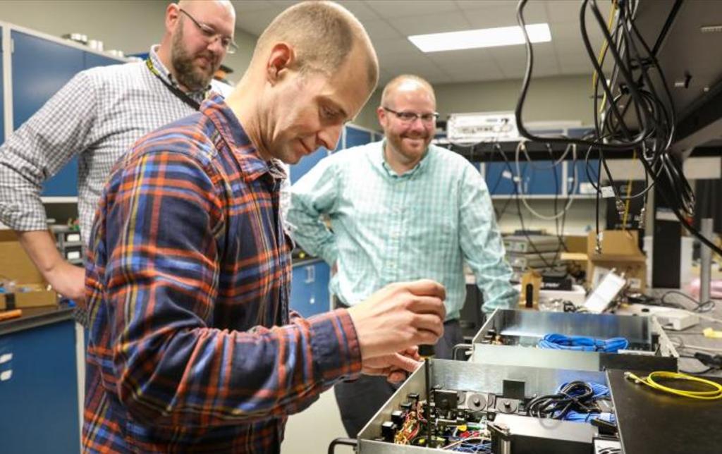 Three researchers stand beside fiber-optic equipment used in an Oak Ridge National Laboratory quantum key distribution system.