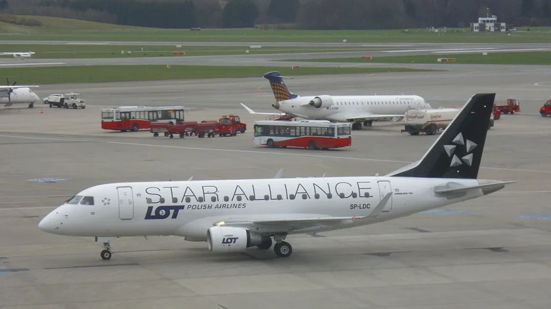 A white LOT Polish Airlines Embraer 170 in Star Alliance livery taxis across the apron at Hamburg Airport.
