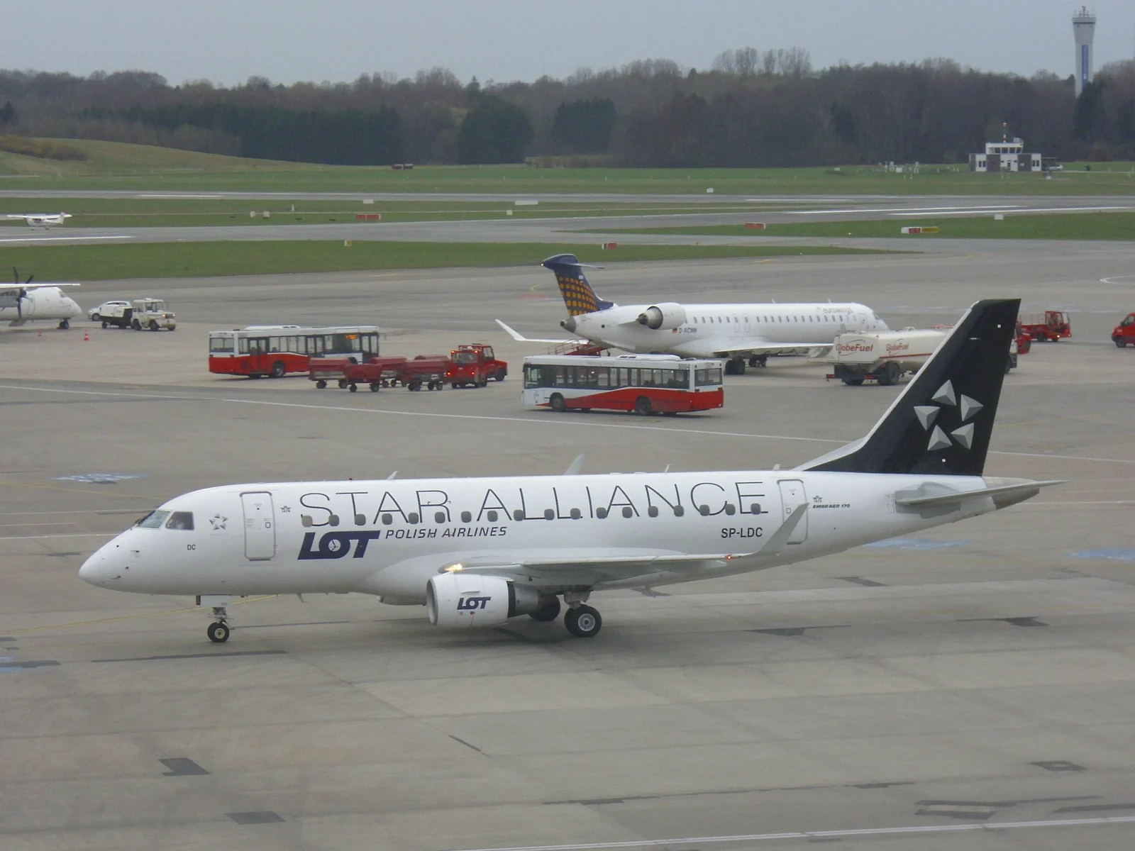 A white LOT Polish Airlines Embraer 170 in Star Alliance livery taxis across the apron at Hamburg Airport.
