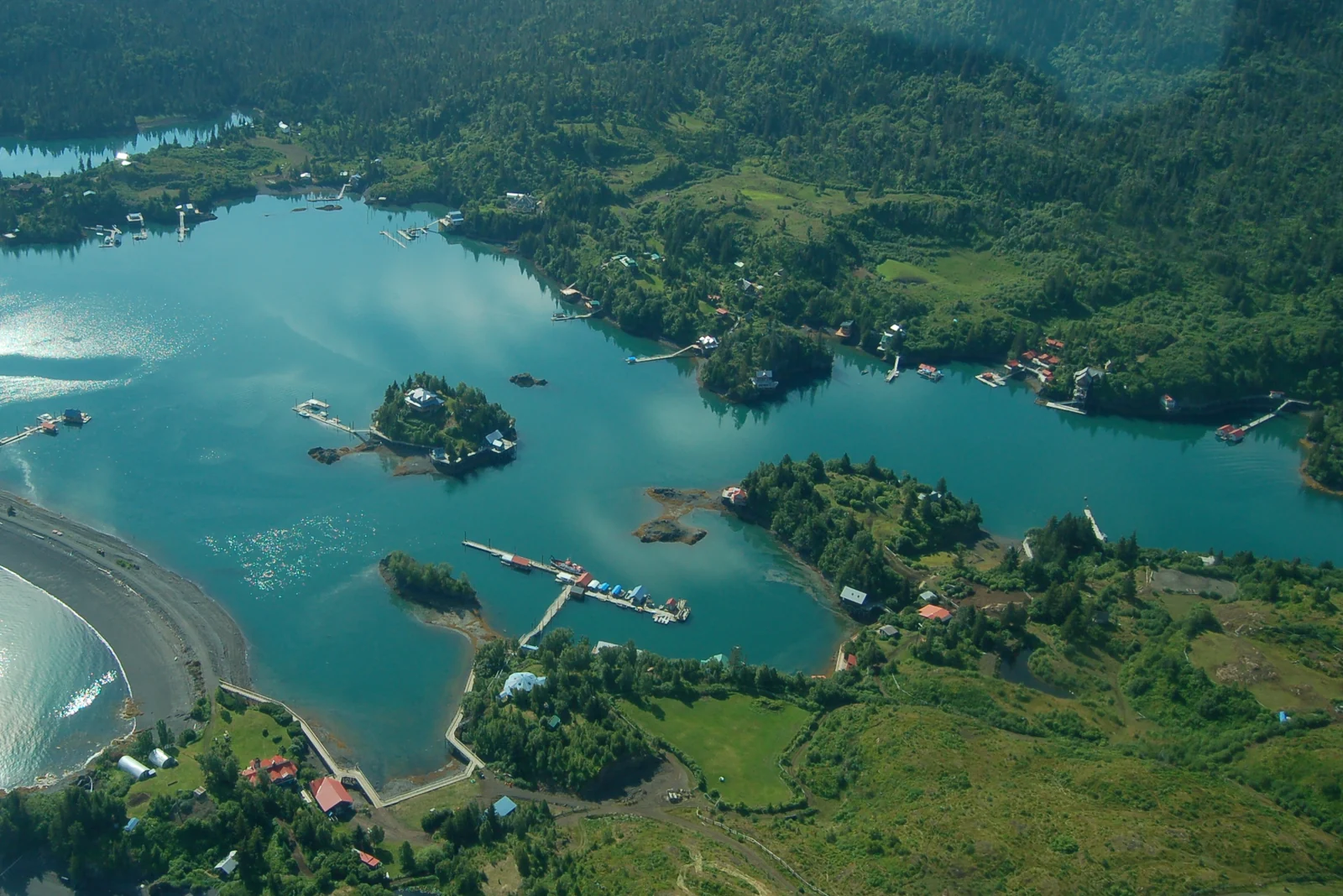 Aerial photograph of turquoise water winding between wooded islands, homes and docks at Halibut Cove, Alaska.