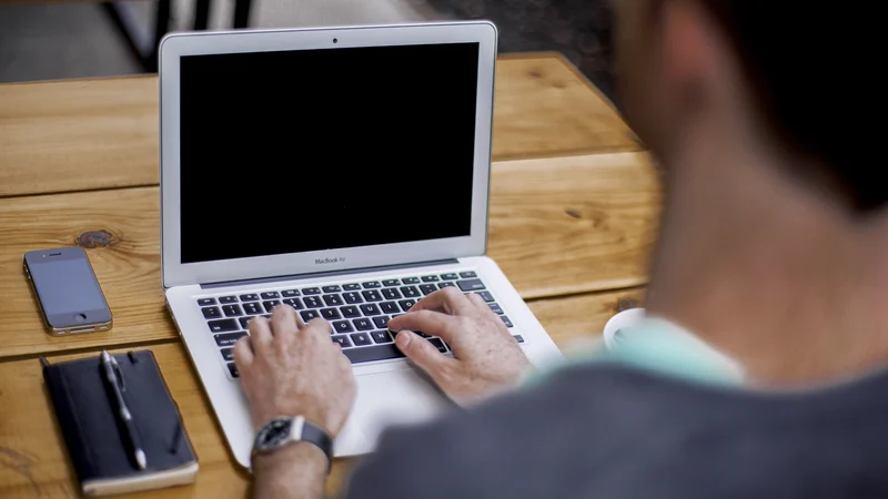 A person typing on a laptop at a wooden table, viewed over their shoulder.