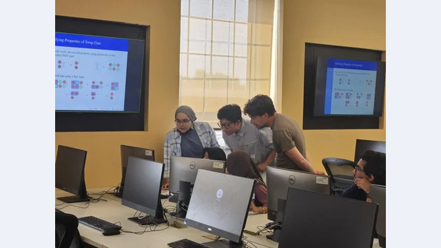 Instructors lean toward participants’ computer screens at a Dalhousie quantum hackathon, with quantum-circuit diagrams projected behind them.