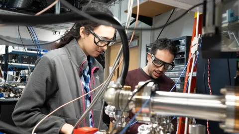 April Sheffield and Baruch Margulis work beside vacuum equipment and control cables in NIST’s Ion Storage lab