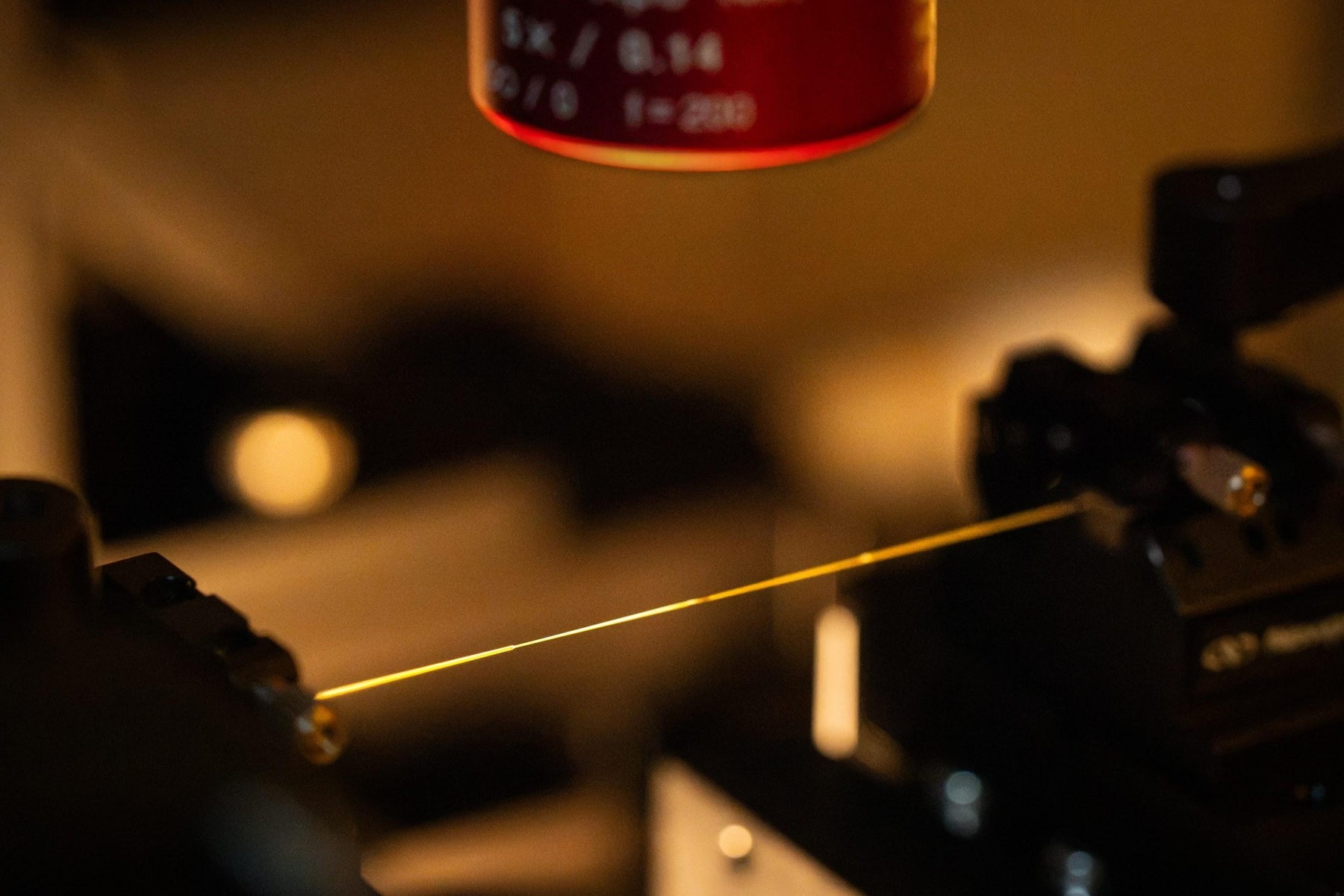A thin glowing optical fiber stretched between laboratory mounts.
