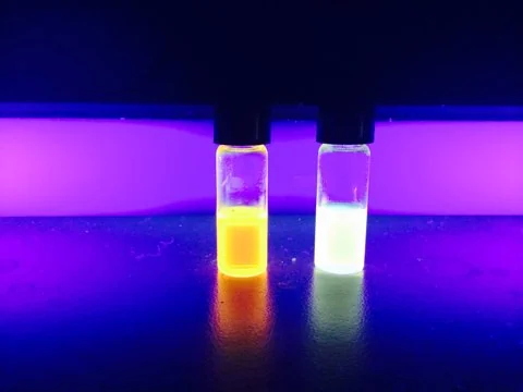 Two vials fluoresce yellow and blue under ultraviolet illumination.