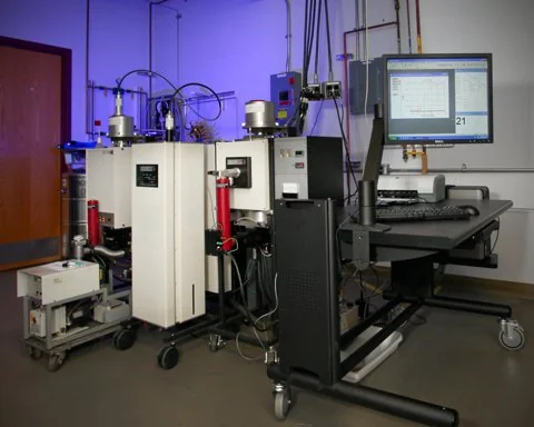 A laboratory instrument stands beside a computer monitor and metal gas lines.