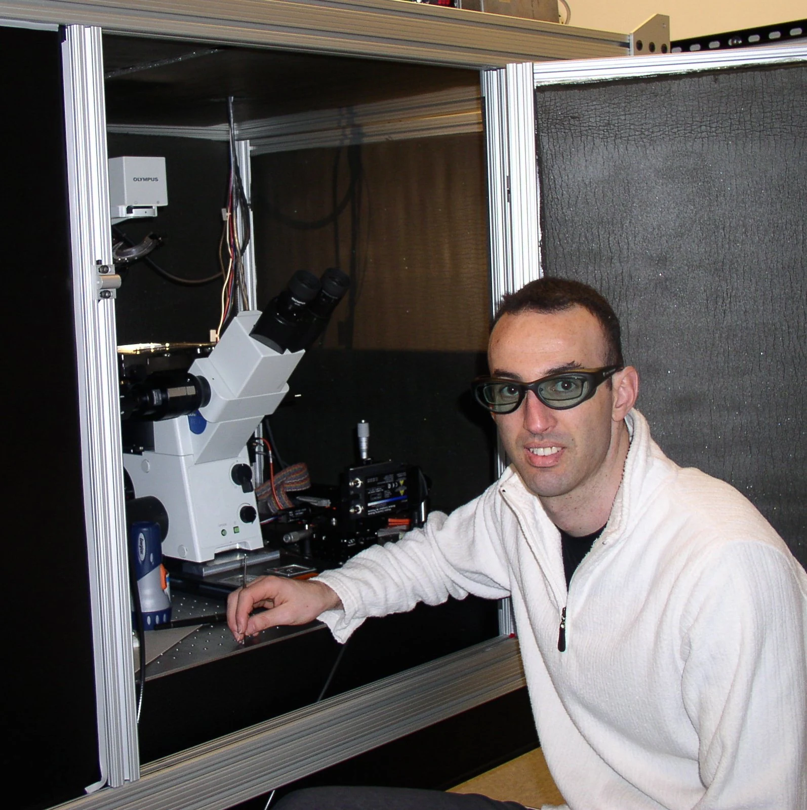 A researcher wearing protective glasses sits beside an optical microscope.