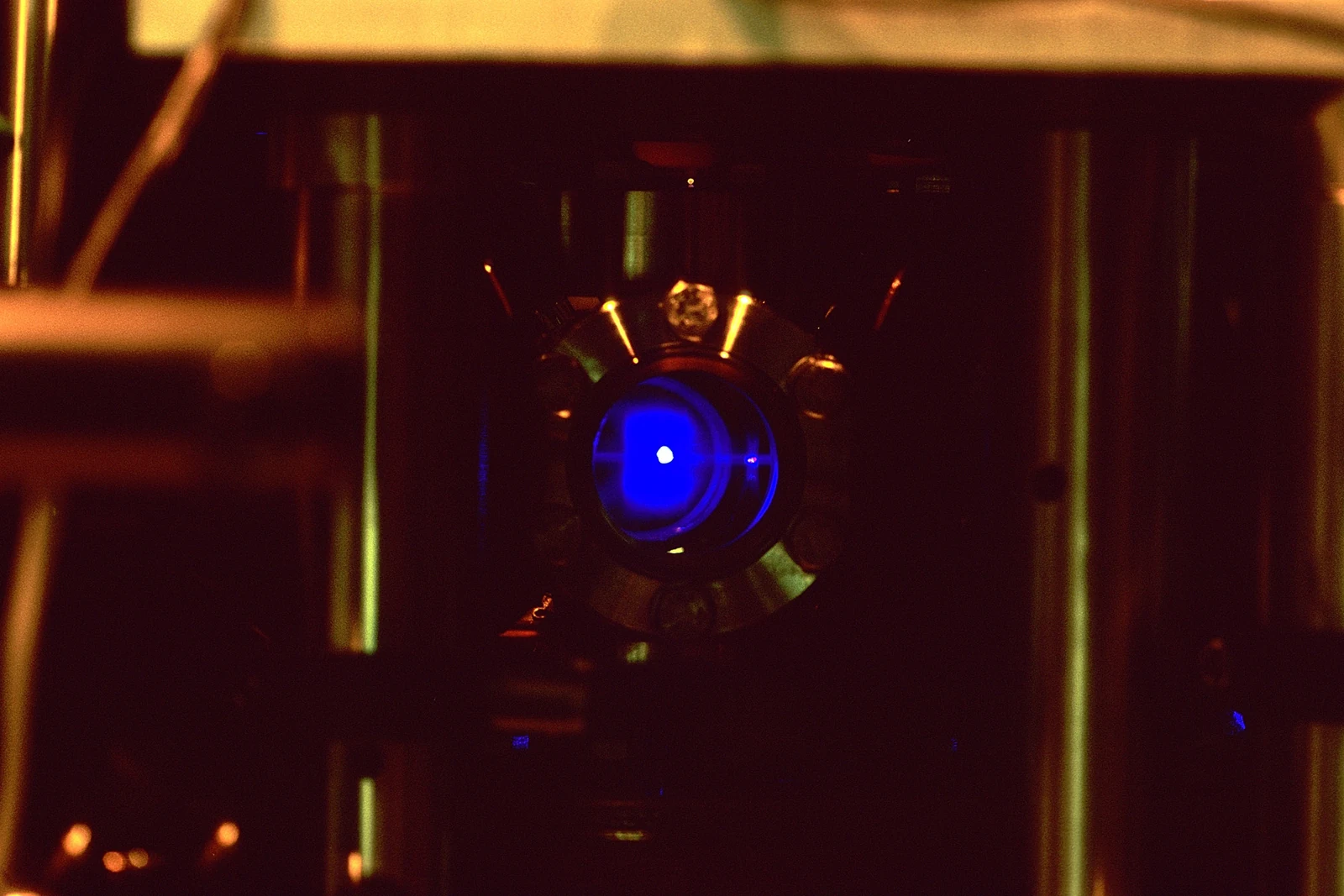Blue fluorescence at the center of a dark vacuum chamber surrounded by optical hardware.