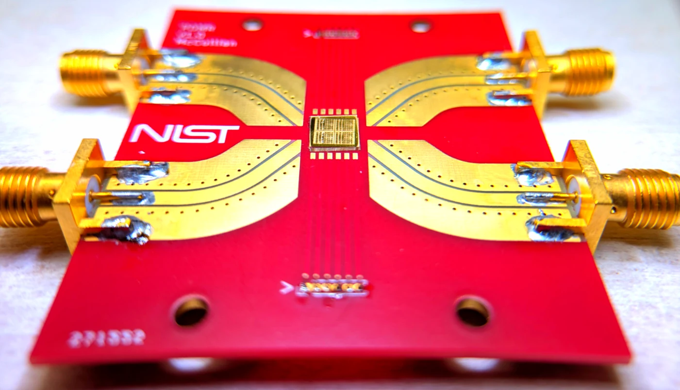 A small gold-colored sensor chip at the center of a red NIST board with gold electrical connectors.