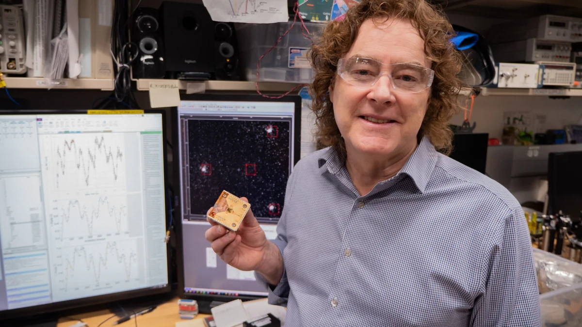 Andrew Wilson holds a small ion-trap chip beside computer displays showing measurement traces and bright trapped atoms in a NIST laboratory.