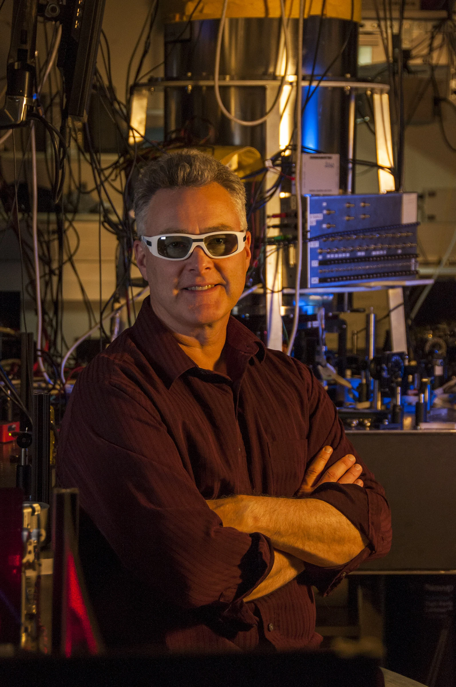 Physicist Andrew Wilson wearing protective glasses in front of a cylindrical vacuum chamber and laboratory instruments.