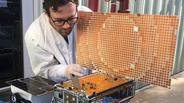 An engineer works beside a small CubeSat with an unfolded square antenna and solar panel.