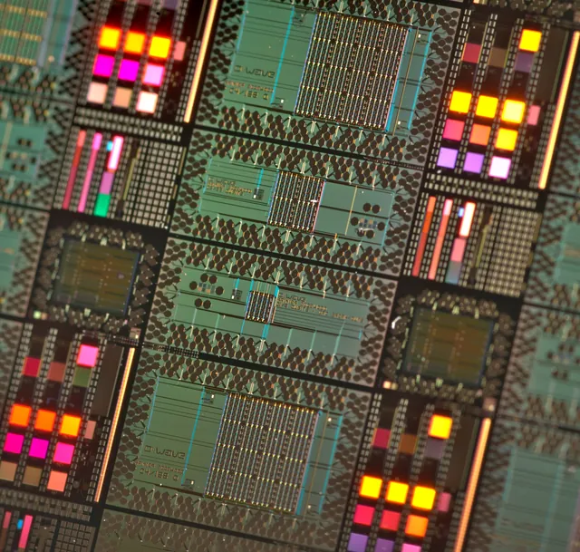 A close view of a patterned D-Wave wafer reflects coloured light from processor regions.