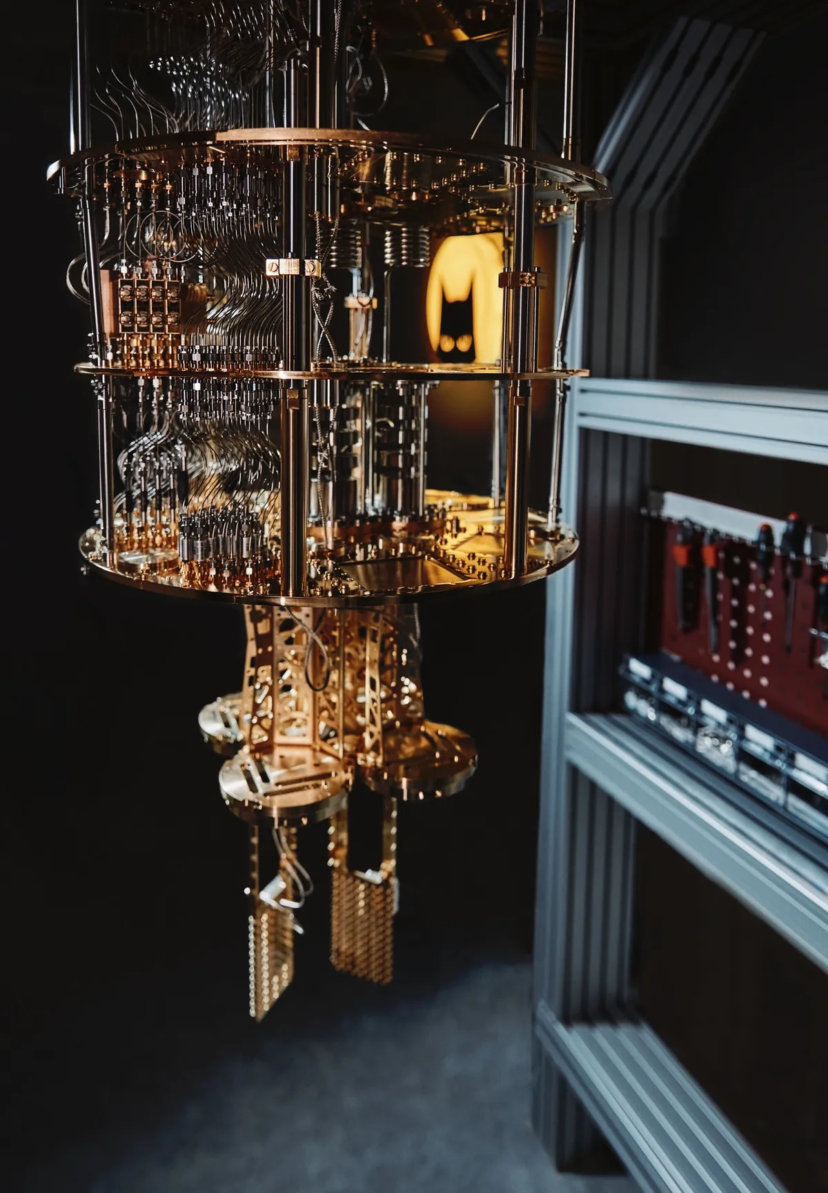 An exposed multi-stage cryostat with dense wiring beside a laboratory equipment rack.