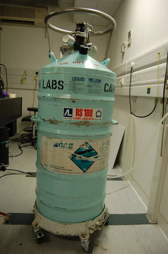 A tall insulated liquid-helium vessel with valves, tubing and safety labels.