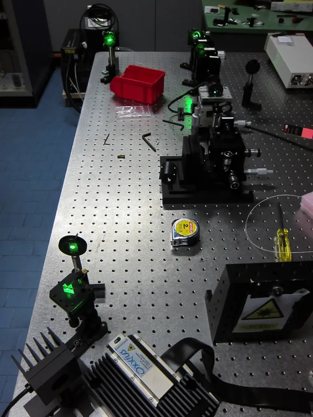 An optical table holds mirrors, mounts and a visible green laser beam.