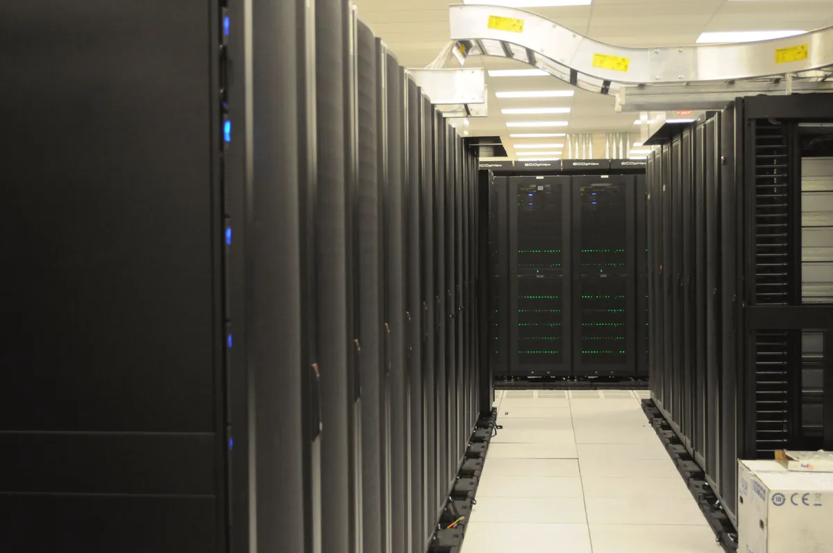 Rows of dark server racks with overhead cable trays in a computing facility.