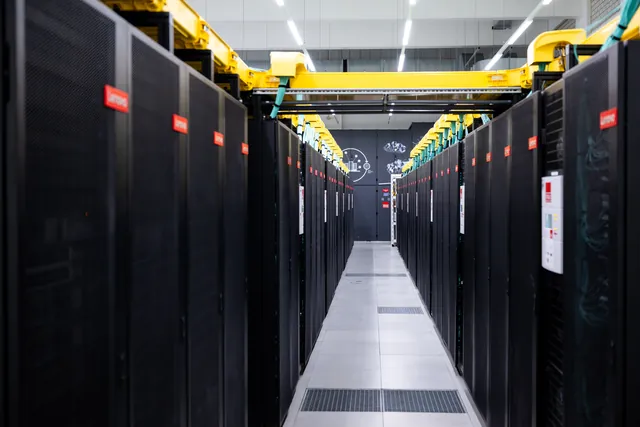 A long aisle of black supercomputer cabinets is topped by yellow cable trays.