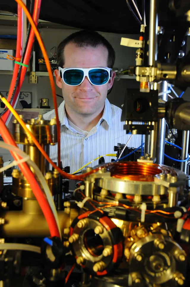 NIST physicist Andrew Ludlow stands beside an optical table filled with atomic-clock equipment.