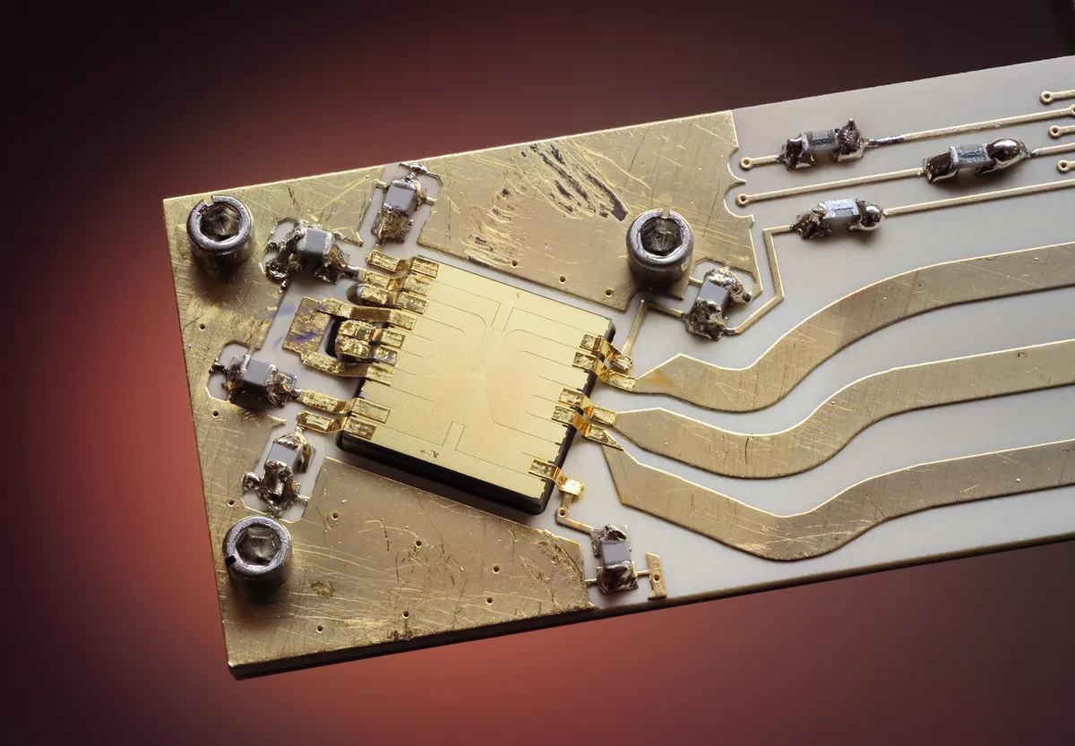 A small gold ion-trap chip sits on a backing with wires and electrical connections.