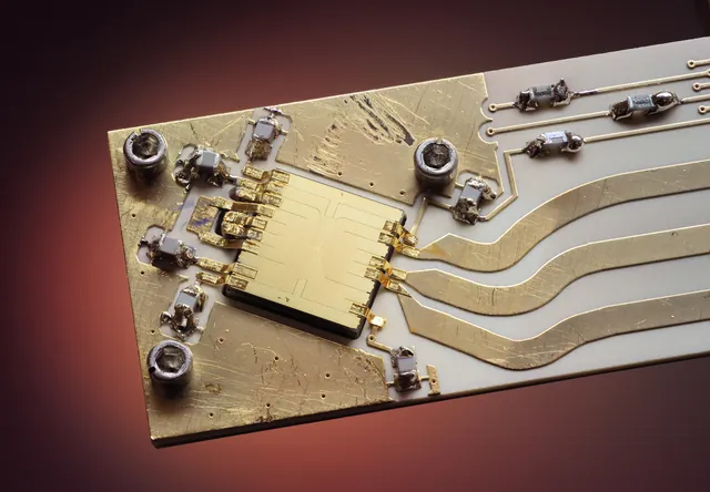 A small gold ion-trap chip sits on a backing with wires and electrical connections.