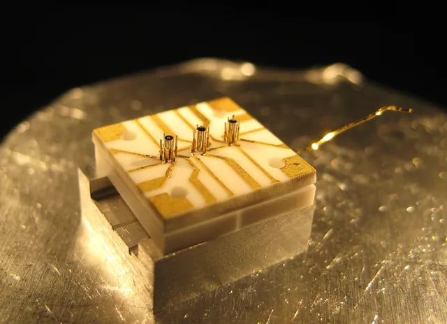A gold microfabricated ion-trap device has three sets of small posts on its surface.
