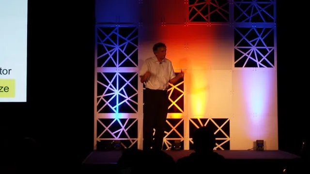 John Martinis speaks on a conference stage beside a projected presentation.