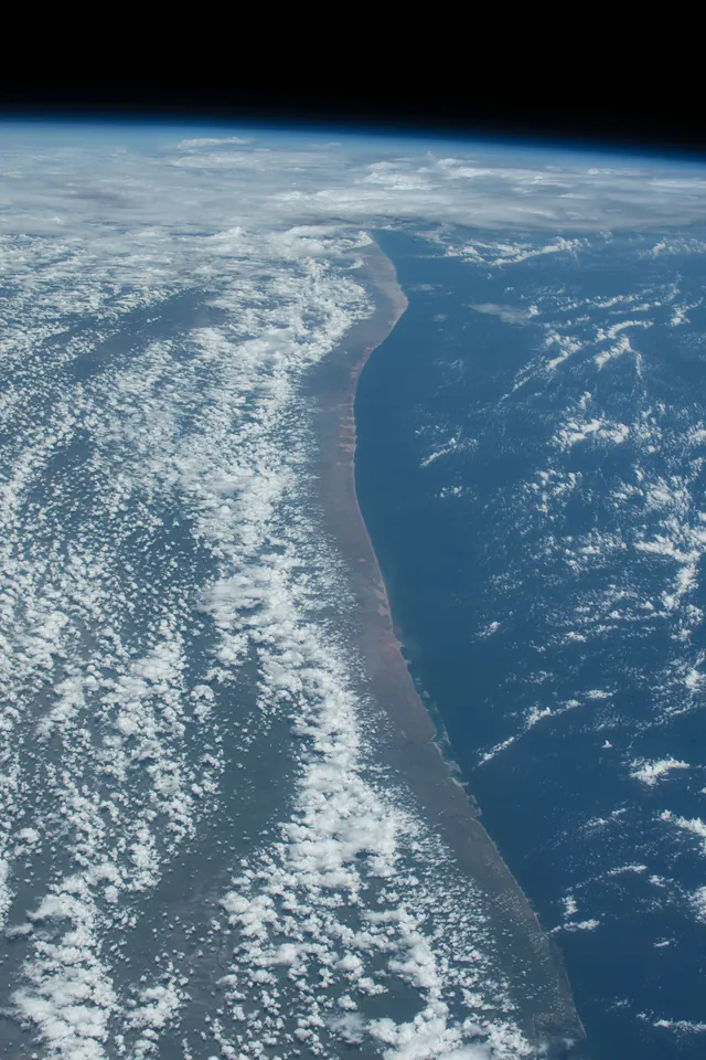 A coastline and banks of clouds curve beneath the dark sky in a view from orbit.