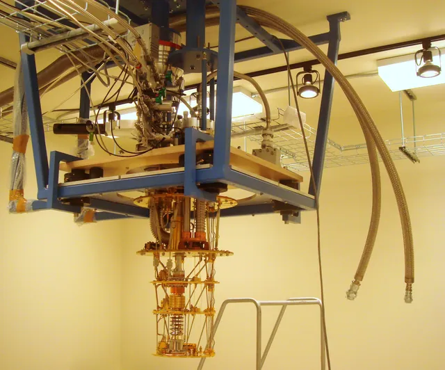 An exposed dilution refrigerator hangs beneath a metal support frame and laboratory tubing.