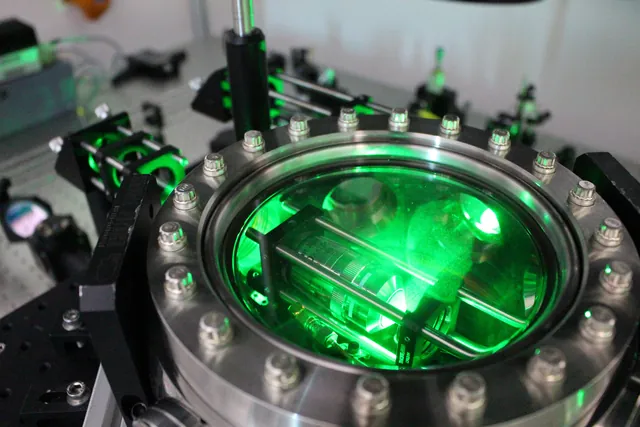 A circular vacuum chamber glows green among lenses and optical mounts on a table.