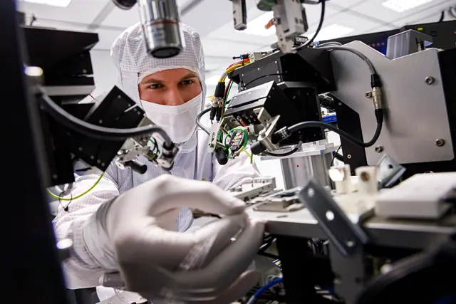 A masked cleanroom researcher examines a small sample under fabrication equipment.