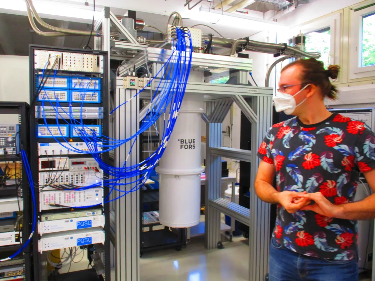 A researcher beside a cryogenic cylinder and racks of laboratory control electronics at Jülich.