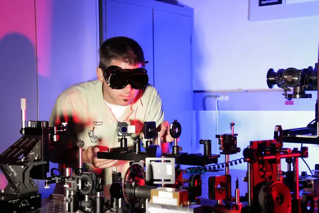 A researcher wearing laser goggles adjusts components on an illuminated optical table.