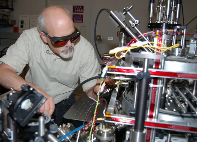 A researcher adjusts a compact ion-source apparatus surrounded by lasers and vacuum equipment.
