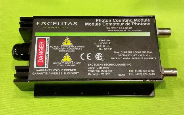 A black single-photon counting module with an optical connection and warning label.