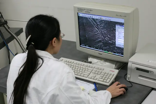 A researcher operates an electron microscope beside a screen showing a magnified sample.