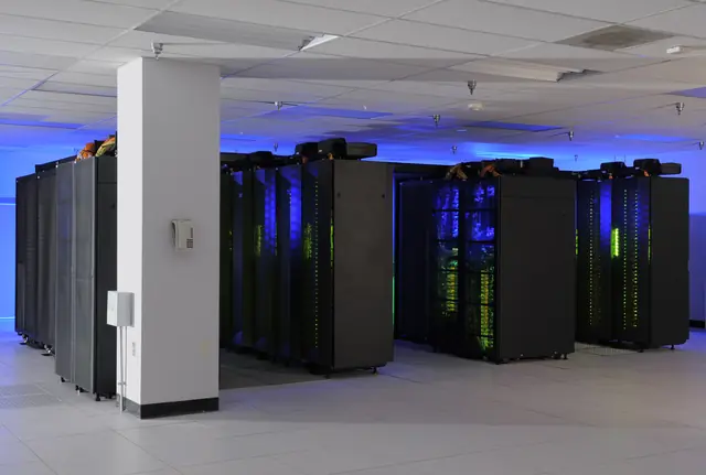 Rows of dark supercomputer cabinets beneath a low white ceiling.