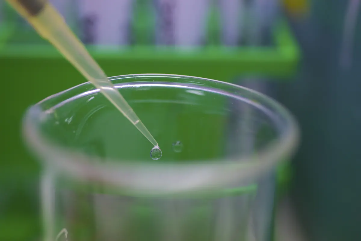 A small liquid drop forms at the end of a pipette above a clear vessel.